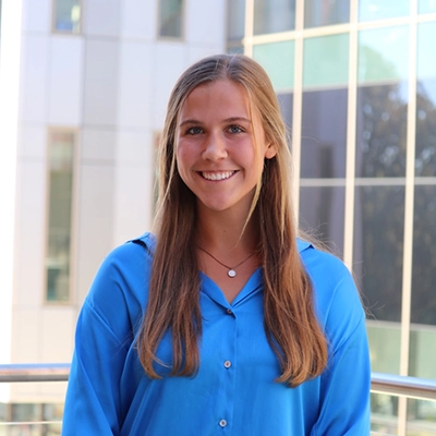 Smiling young woman with long hair wearing a blue shirt, standing in front of a modern building.