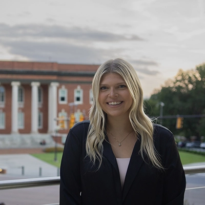 A smiling woman stands outdoors in front of a university building during sunset.