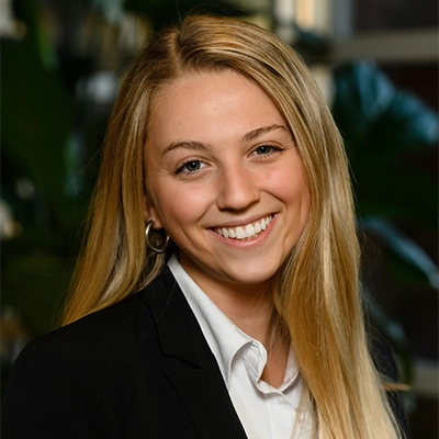 Professional headshot of a young woman with long blonde hair, wearing a black suit jacket and a white shirt, smiling against a backdrop of greenery.