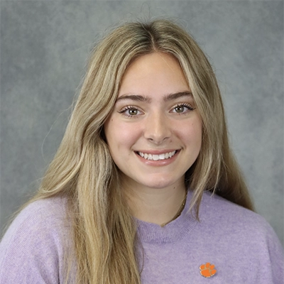 Headshot of a young woman with long blonde hair, wearing a light purple sweater and an orange Clemson University logo pin.