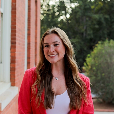 A young woman in a pink blazer smiles confidently in front of a red brick building and greenery.