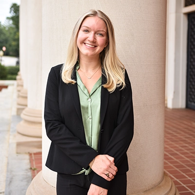 Professional portrait of a woman in a black blazer and green blouse, smiling in front of large columns.