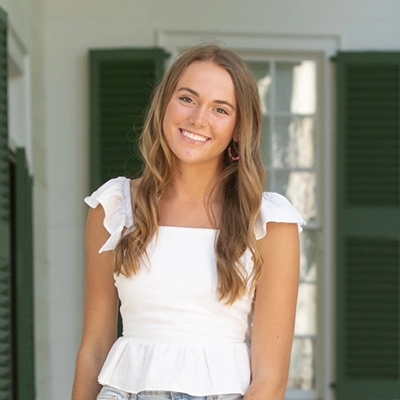 A young woman with long hair smiling, wearing a white peplum top with ruffled sleeves, standing in front of green shutters.