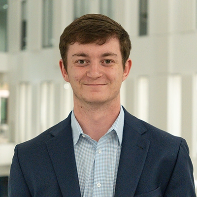 Professional headshot of a young man in a blazer, smiling in a modern indoor setting.