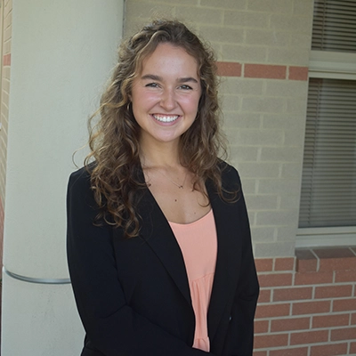 A smiling young woman with curly hair wearing a blazer and a light-colored top, posing confidently in an outdoor setting.