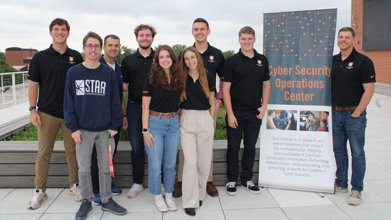 A group of students, CISO John Hoyt and CSOC Manager Calvin Watts stand on the roof of the Watt Center with a sign that says Cybersecurity Operations Center.