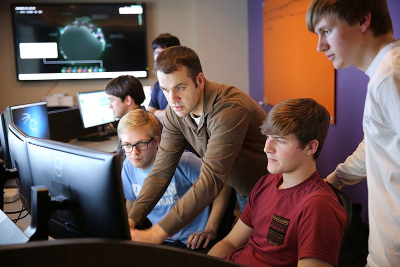 Multiple people sitting in a room with lots of desktop computer with one person pointing at the screen of one computer showing another person something.