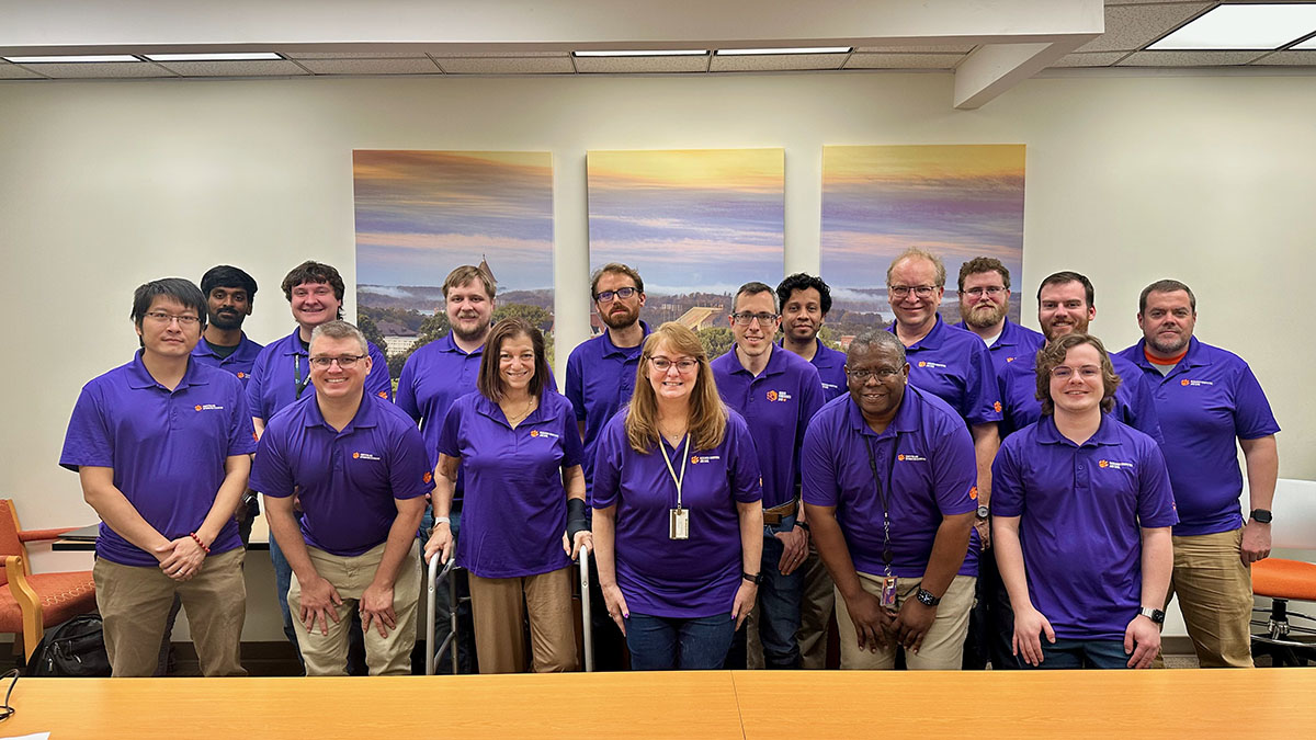 Clemson Research Computing and Data Staff standing together wearing purple polos with the RCD logo on it smiling in a conference room.
