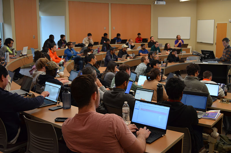 A presentation setup in a classroom, featuring a speaker explaining a topic, with students attentively listening and using laptops.