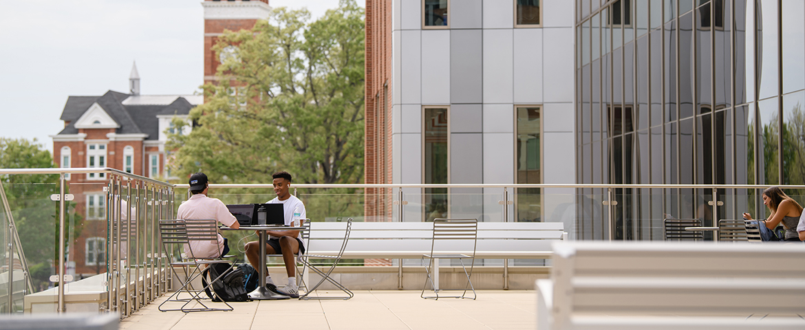 Two students sitting at a table with each other and working on their laptops with trees and a red brick building (tillman Hall) in the background.