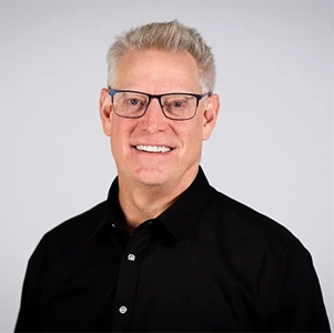 Portrait of a smiling middle-aged man with glasses, wearing a black shirt against a neutral background.