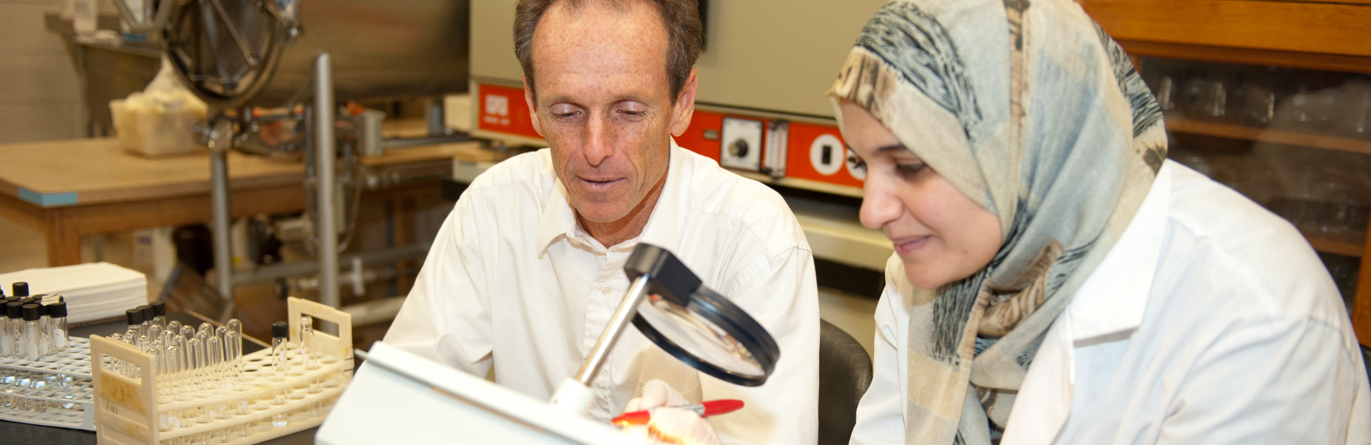 Male and female wearing gloves and looking through magnifying glass making notes while conducting research.