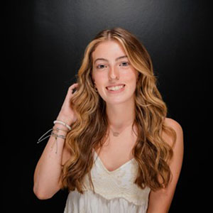 Smiling young woman with long wavy hair in a white sleeveless top against a dark studio background.
