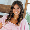 Smiling woman with long wavy hair wearing a light pink top indoors.