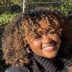 Smiling woman with curly hair wearing a green blouse against a gray studio backdrop.