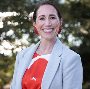 Smiling woman in a grey blazer and a red and white dress, standing outdoors with a blurred natural background.