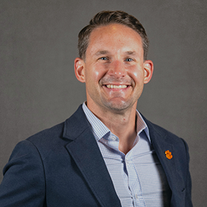 Professional headshot of a smiling man in a dark blazer with a light blue shirt, featuring a Clemson University logo pin.