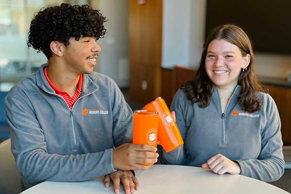 Two Honors students sitting at a table in the Great Hall, and smiling as they clink two Honors-branded cups together.