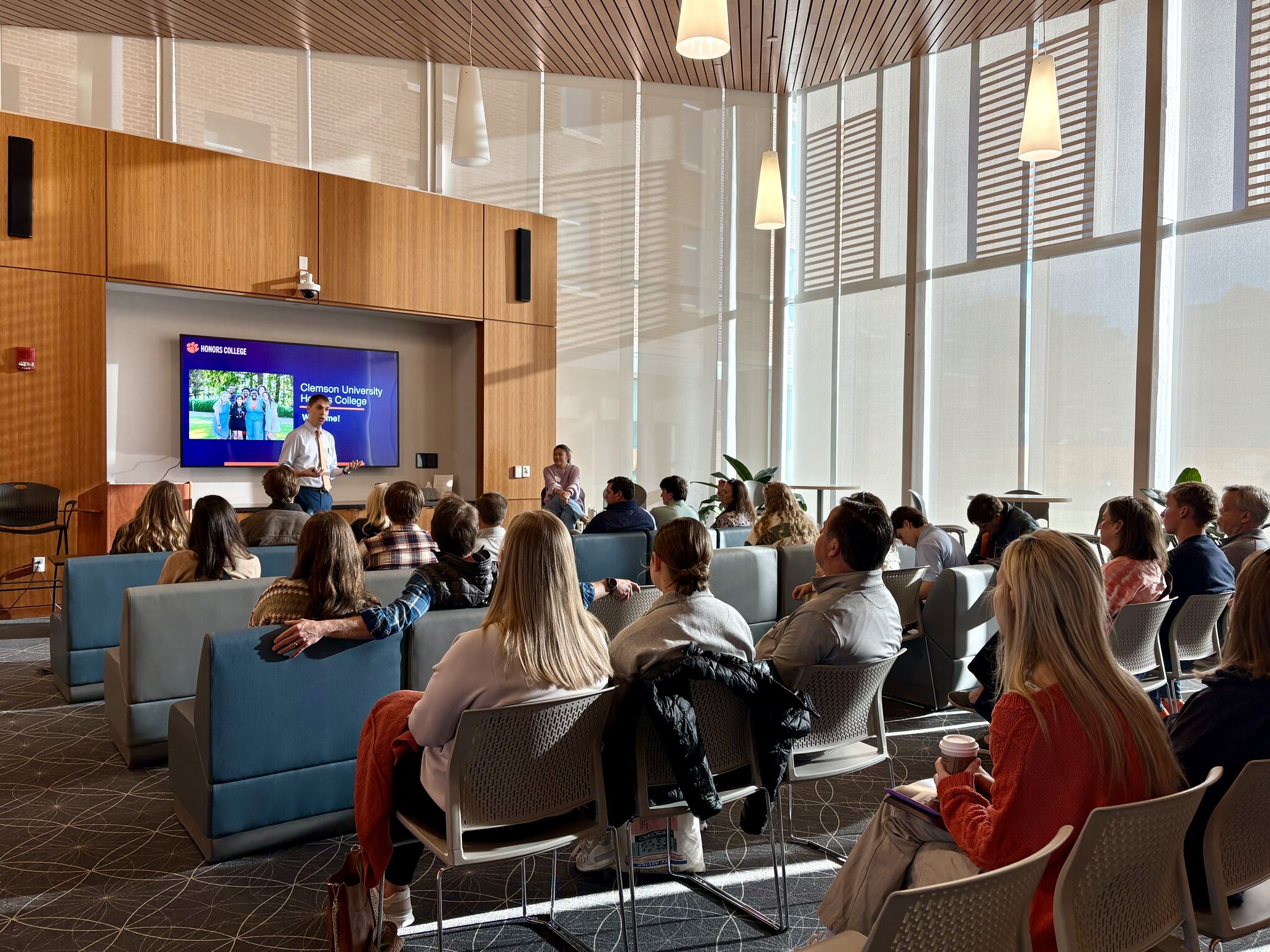 A group of people sitting in the Honors College's Great Hall, theatre-style. A man is speaking at the front of the room beside a presentation screen.