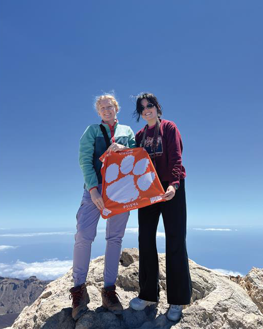Two women standing on top of a clifftop with their backs to the ocean, holding a Clemson Tiger Rag.