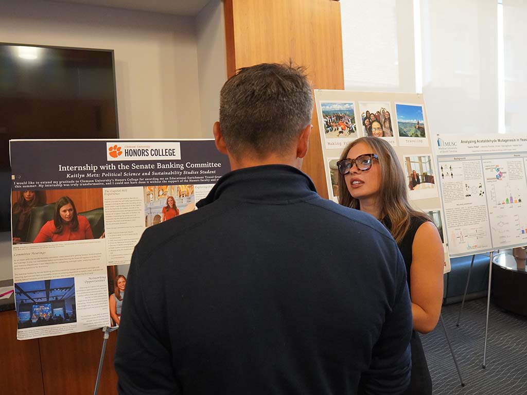 An Honors student talking to a man standing with his back to the camera. They are standing in the Honors College Great Hall and in front of the student's poster.