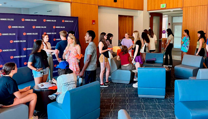 Several Honors students and families mingling together in the Great Hall in the Honors Center on campus. A family is talking to an Honors advisor in front of a large Honors College backdrop.