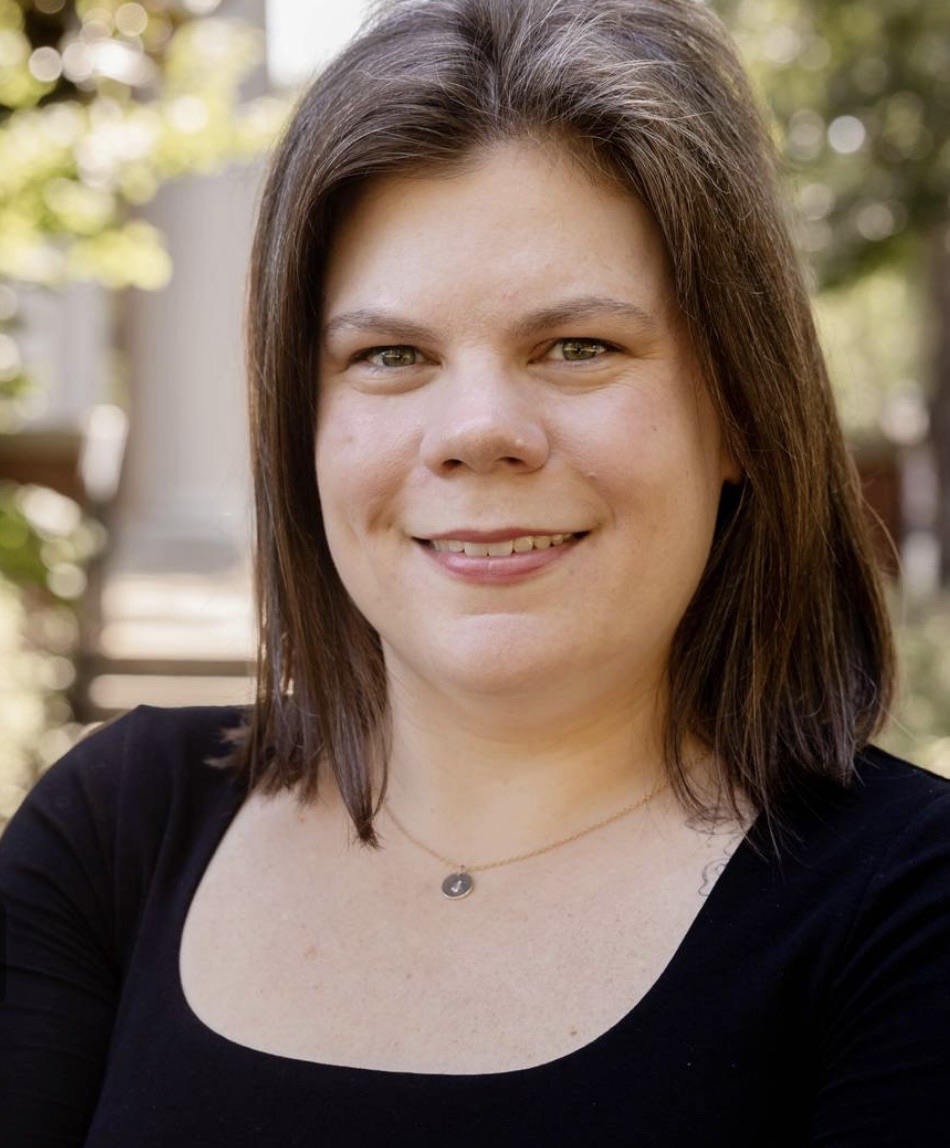 Professional headshot of a woman with shoulder-length brown hair, wearing a black top and smiling outdoors.