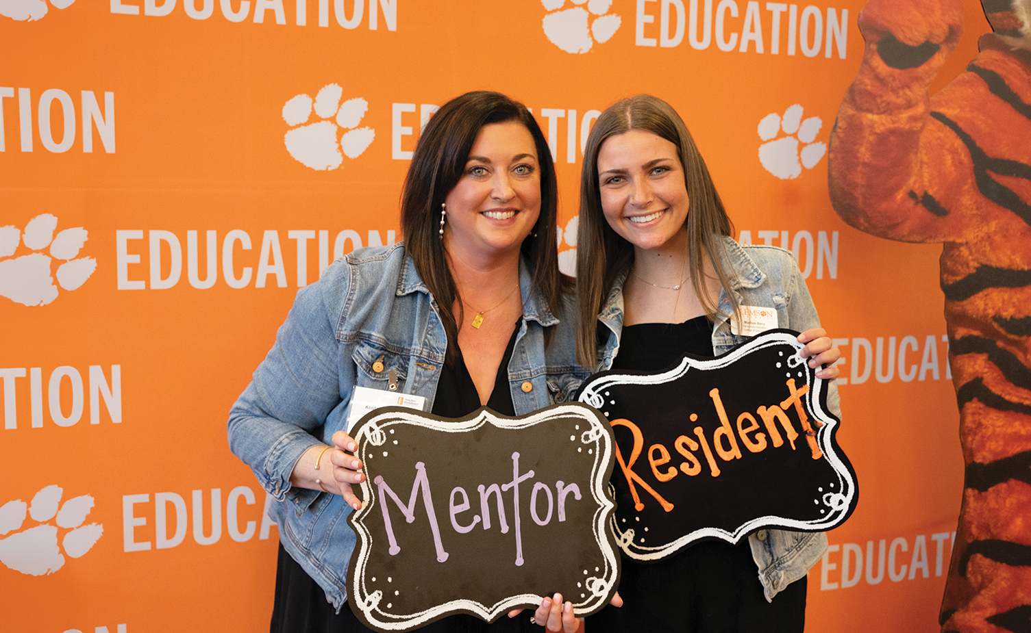 Two women smiling and posing with signs that read "Mentor" and "Resident" in front of an orange backdrop with paw print designs and the word "EDUCATION."