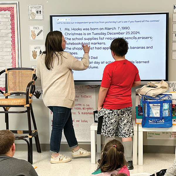 A teacher and student engage in a lesson at the front of a classroom, with other students attentively seated nearby.