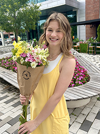 A woman in a yellow dress holds a bouquet of colorful flowers while smiling in an outdoor setting.