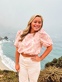 A young woman smiling while standing on a coastal cliff with ocean views, wearing a pink floral top and white pants.