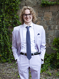 A young man in a light purple suit and glasses stands confidently against a stone wall, surrounded by greenery.