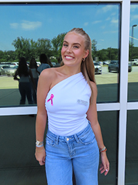 A young woman smiles while wearing a one-shoulder white top with a pink ribbon, paired with high-waisted jeans, standing in front of a glass window.