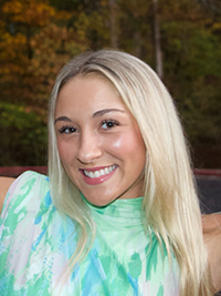 Smiling young woman wearing a colorful, tie-dye blouse, set against a blurred autumn background.