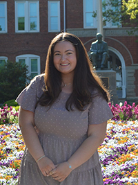 A smiling young woman stands in front of a colorful flower bed and a statue on a university campus.