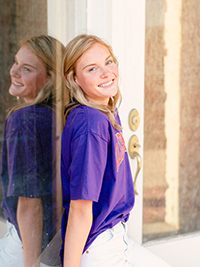 A smiling young woman in a purple t-shirt leaning against a glass door, reflecting a sense of happiness and casual style.