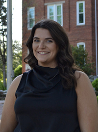 Woman with a dark, sleeveless blouse stands in front of a brick academic building.