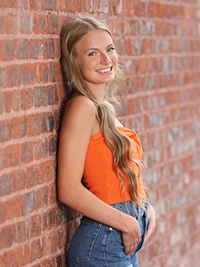 A young woman in an orange top poses confidently against a brick wall.