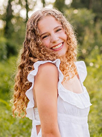 A smiling young woman with long, curly hair, standing outdoors amidst greenery, wearing a white dress with ruffled sleeves.