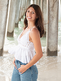 A young woman smiling while posing under a pier, wearing a white top and blue jeans.