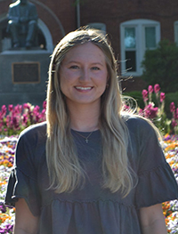 A young woman with long blonde hair stands in front of a colorful flower garden, smiling warmly.