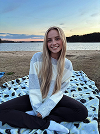 A young woman sitting on a blanket by a lake at sunset, smiling while wearing a cozy striped sweater.