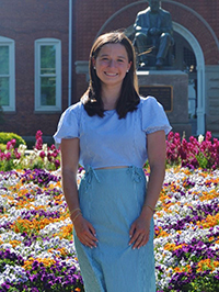 A smiling woman stands in front of a colorful flower garden with a brick building in the background.