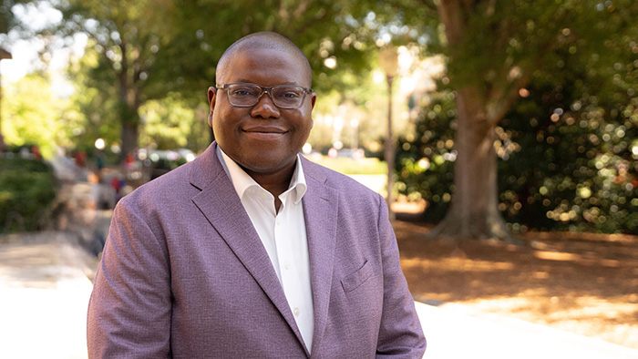 A man with glasses smiles while wearing a purple suit jacket and a white collared shirt. He stands outdoors surrounded by trees and green leaves, captured in bright midday light.