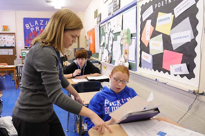 A teacher assists a student in a classroom setting, while another student works quietly in the background.