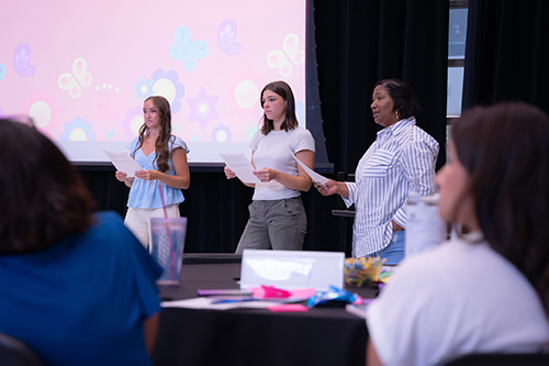 Three women presenting at an event, with a colorful backdrop featuring butterflies and flowers.