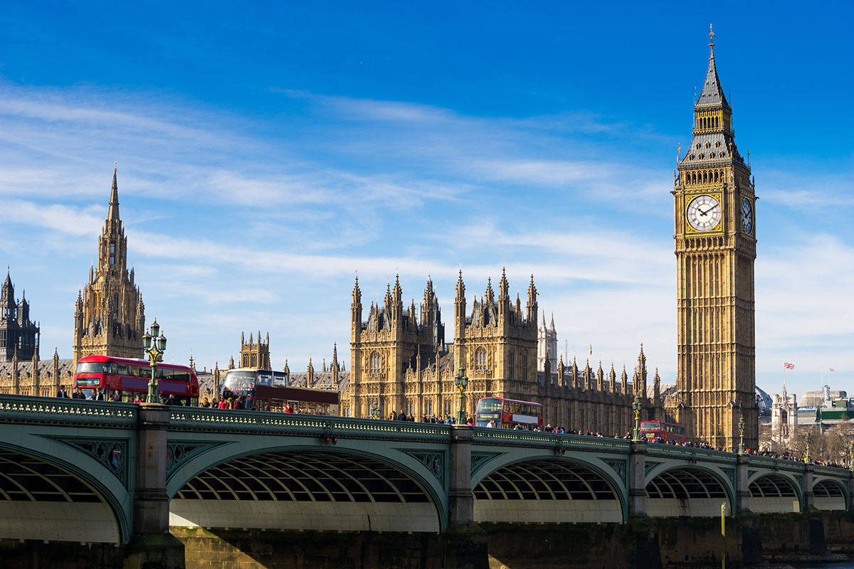 The iconic Big Ben clock tower and the Houses of Parliament along the River Thames in London, featuring red double-decker buses crossing the bridge.