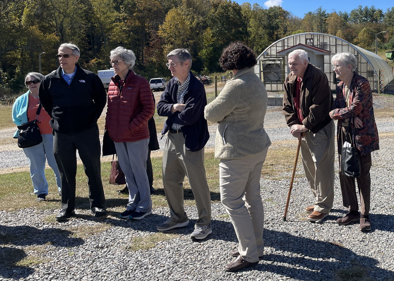 A group listens to a speaker as they stand outdoors with a greenhouse in the background