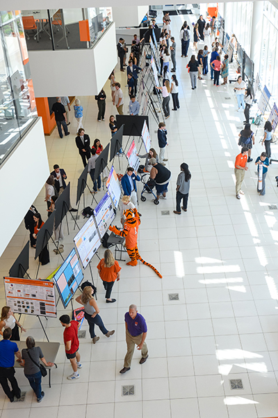 A bustling event scene showcasing attendees interacting with displays, featuring a person dressed in a tiger costume among the crowd.