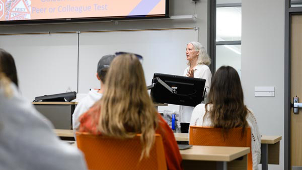 an older woman speaks to a group of students from the front of a classroom